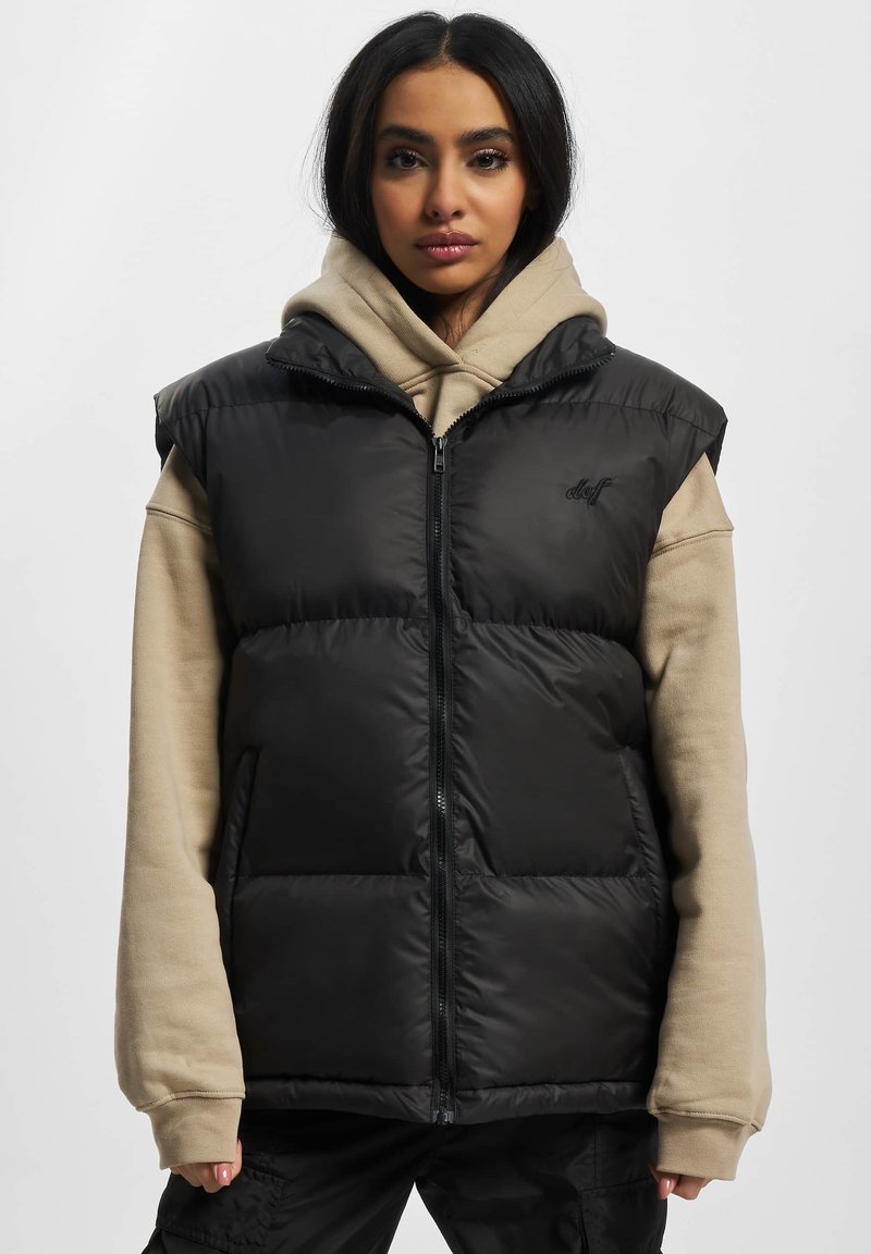 Schwarze wärmende Weste ohne Ärmel mit Reißverschluss, zwei Seitentaschen und glänzendem Finish, darüber getragen einen beigen Hoodie.