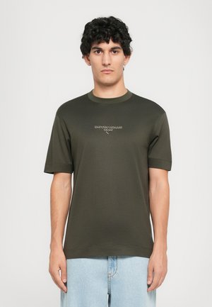 Young man with curly dark hair wearing a dark green Emporio Armani Milano t-shirt and light blue jeans, standing against a plain background.