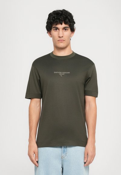 Joven con pelo rizado oscuro, vistiendo una camiseta de Emporio Armani Milano en color verde oscuro y jeans azul claro, de pie contra un fondo sencillo.