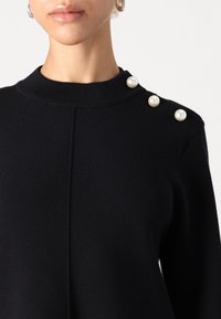 Närbild av kvinna som bär en svart tröja med tre stora pärlknappar på vänster axel och en liten guldring som örhänge.