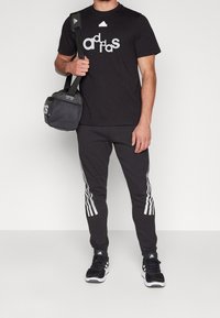 Svart träningströja med vit "adidas"-logotyp, svarta träningsbyxor med vita sidsrum, och svarta sneakers. Bär en svart gymväska.