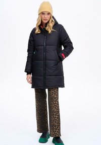 Čierna puffrová bunda s kapucňou, so zipsom na prednej strane, dvoma bočnými vreckami a červenými akcentmi. Nosí sa s nohavicami s leopardím vzorom a zelenými topánkami.