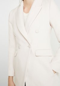 Blazer de lana rosa claro con diseño de doble botonadura, botones redondos y bolsillos laterales. Presenta un cuello de solapa y un ajuste entallado.