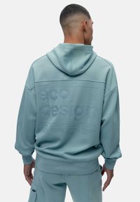 Pehmea sinine kapuutsiga dressipluus, mille seljal on tekstureeritud "ökodisaini" sõnum, lõdva lõikega ning ribidega varrukate ja äärtega.