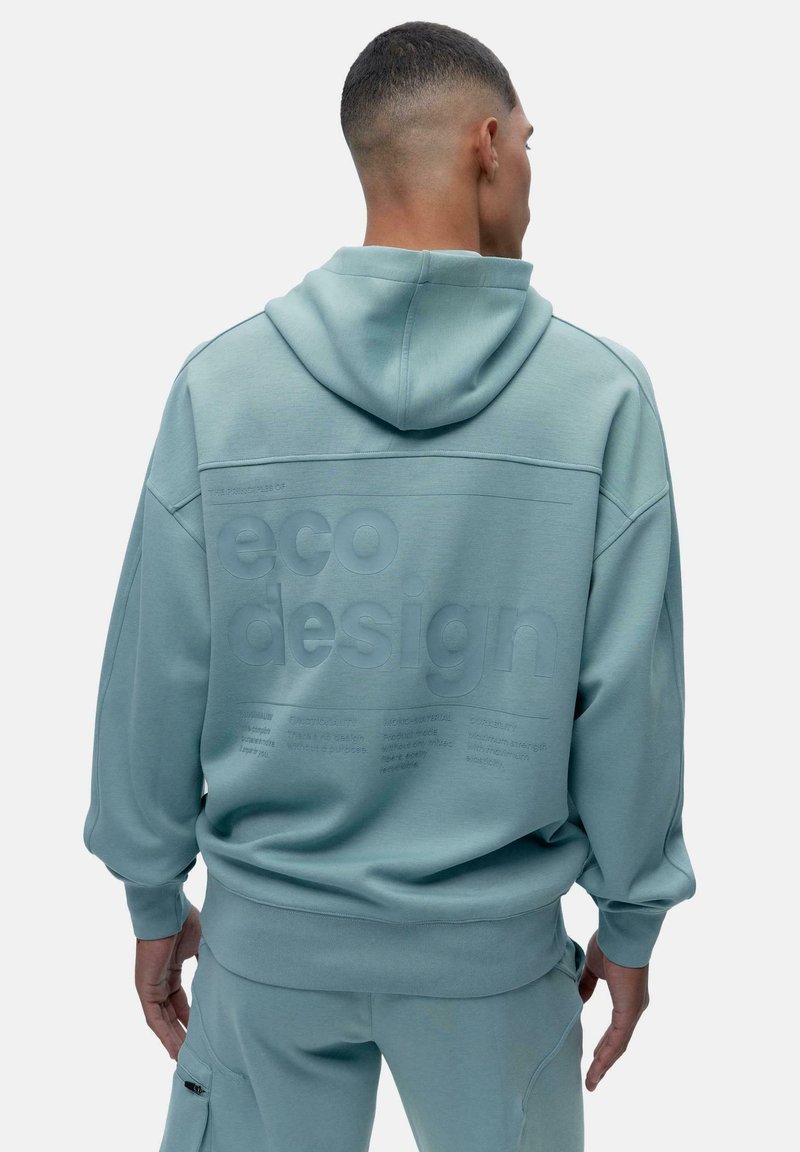 Pehmea sinine kapuutsiga dressipluus, mille seljal on tekstureeritud "ökodisaini" sõnum, lõdva lõikega ning ribidega varrukate ja äärtega.