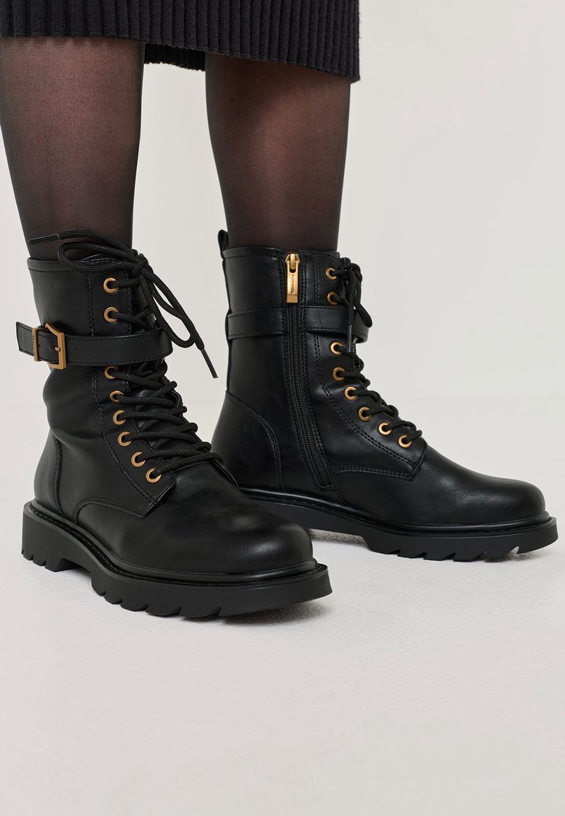 Bottes courtes en cuir noir avec semelles épaisses, lacets, fermetures éclair latérales et détail de boucle dorée, portées avec des collants noirs transparents.