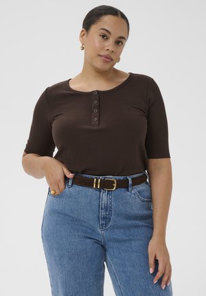 Žena u smeđoj kratkoj majici s dugmićima, plavim trapericama visokog struka, tamno smeđim remenom i zlatnim naušnicama u obliku obruča.