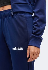 Άτομο που φοράει σκούρο μπλε φόρμα Adidas, εμφανίζοντας το λογότυπο της μάρκας στο αριστερό μηρό και έχοντας το χέρι χαλαρό στο πλάι.
