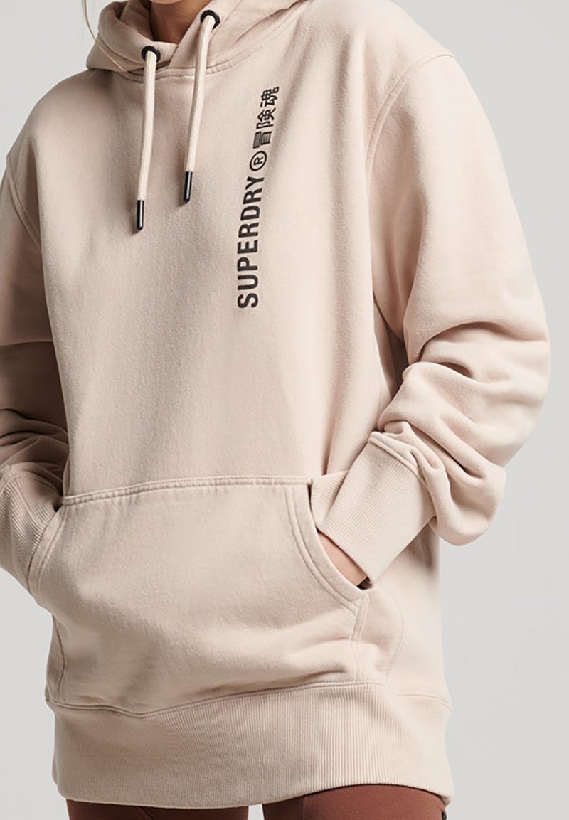 Persoon draagt een beige Superdry-hoodie met verticale zwarte logotekst, handen in de grote voorkantzak, tegen een effen lichte achtergrond.