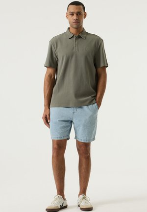 Polo vert olive avec un tissu texturé, patte à deux boutons ; associé à un short en denim bleu clair. Des baskets blanches décontractées complètent le look.