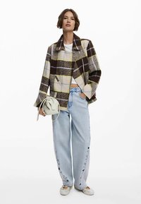 Giacca a quadri marrone e crema con colletto a risvolto, abbinata a jeans a gamba larga azzurri, con una piccola borsa beige in mano. Scarpe basse completano il look.