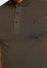 Tmavě olivově zelené polo tričko vyrobené z hladké bavlněné tkaniny, s límečkem, tříknoflíkovou légou a malou nášivkou s logem.