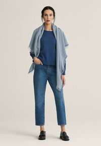 Sininen neule, jonka päällä on vaalea sininen frillapusero, yhdistetty suoriin farkkuihin ja mustiin loafer-kenkiin neutraalin taustan kanssa.