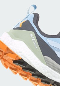 Chaussure de trail running avec une tige gris foncé, des accents bleu clair, des surpiqûres texturées, une semelle intermédiaire blanche amortie et une semelle extérieure orange adhérente.