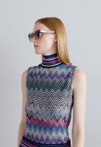 Débardeur en tricot sans manches avec un col haut, présentant un motif en chevrons dans des tons de noir, bleu, violet et rose. Accompagné de lunettes de soleil foncées.