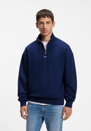 Junger Mann trägt einen dunkelblauen Sweatshirt mit Reißverschluss über einem weißen Hemd und hellblauen Jeans vor einem schlichten weißen Hintergrund.