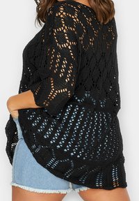 Svart stickad tröja med en öppen vävstruktur, 3/4-långa ärmar och en lös passform, kombinerad med ljusblå denimshorts.