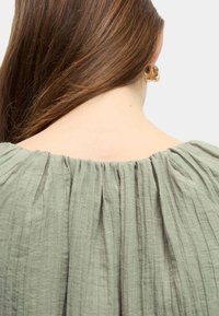 Top verde plissettato con scollatura ampia, tessuto leggero e arricciature sul retro. I dettagli notevoli includono cuciture testurizzate e un drappeggio morbido.