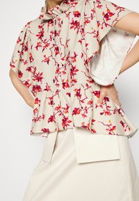 Donna che indossa una blusa floreale beige, rossa e arancione con maniche a sbuffo e orlo arricciato, abbinata a una gonna leggera beige con tasche a sovrapposizione.