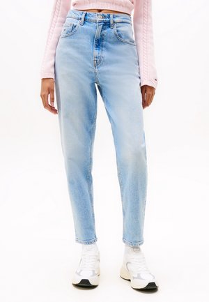 Persoon draagt lichtblauwe high-waist jeans, roze cropped trui en witte sneakers, staand tegen een witte achtergrond.