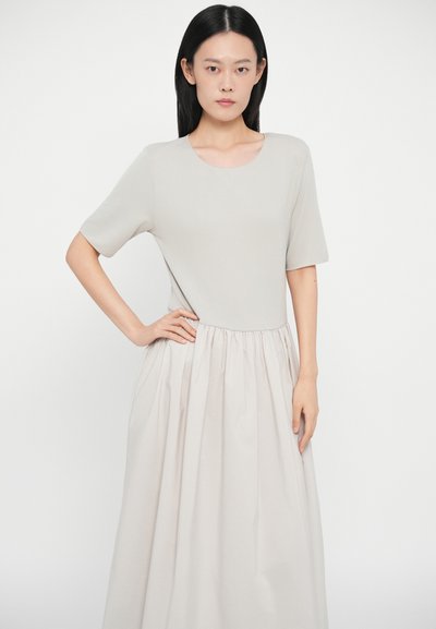 Šviesiai pilka suknelė su prigludusia liemene, trumpomis rankovėmis ir raukšlėta, pilna sijonu. Lygus audinys su švelnia tekstūra, tvarkingos linijos.