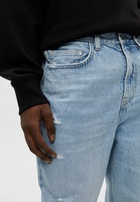 Mano di carnagione scura appoggiata su jeans in denim strappati di un blu chiaro, abbinata a una maglietta nera a maniche lunghe.