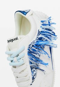 Sneaker bianco con accenti in tessuto frangiato blu e bianco. Caratterizzato da lacci blu e branding sul tallone. Punta arrotondata e suola testurizzata.