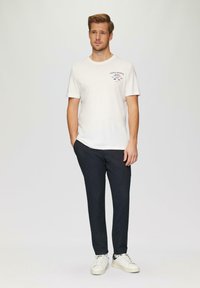 Witte katoenen t-shirt met marineblauwe grafische print op de linkerborst, gecombineerd met donkere broek en witte sneakers, staand tegen een lichte achtergrond.