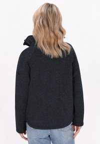 Fleece-takki tummansinisenä, jossa on teksturoitu pinta, täysivetoketju, korkea kaulus ja pitkät hihat. Näkyvää ommeltua saumaa ja kaareva helma takana.