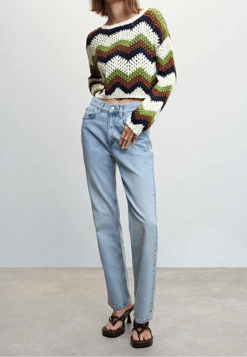 Femme portant un pull court en maille multicolore à motif zigzag, un jean taille haute bleu clair et des sandales à talons noires, debout à l'intérieur.