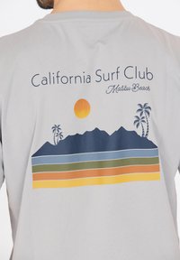 Vaaleanharmaa t-paita, jossa on graafinen kuvasto, jossa on vuoria, aurinko, palmuja ja kerrostettuja raitoja sinisessä, vihreässä, oranssissa ja keltaisessa.