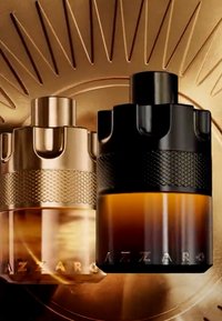 To Azzaro parfume flasker, en guldfarvet og en sort, med teksturerede bånd, placeret mod en gylden solstrålebagsgrund.