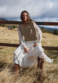 Braune Strickweste mit Blumenstickerei, transparente weiße Ärmel, mehrlagiger weißer Rock und braune kniehohe Stiefel vor natürlichem Hintergrund.