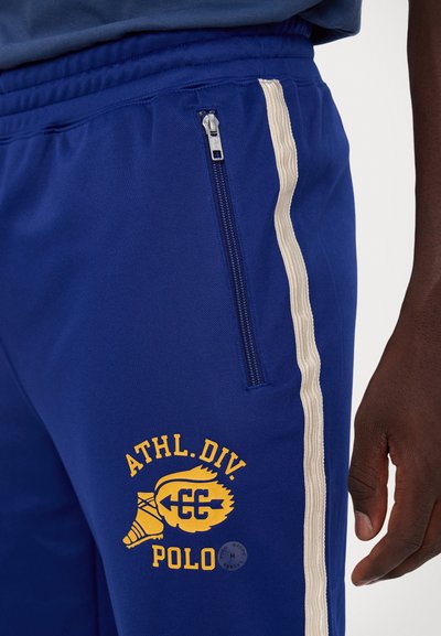 Pantaloni sportivi blu con tasca con zip, striscia laterale crema e grafica gialla "ATHL.DIV. POLO". Realizzati in tessuto morbido e leggero.
