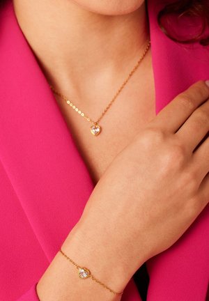 Un collier et un bracelet en or présentent des charms en forme de cœur ornés de pierres claires. Ils sont portés sur un chemisier rose. La chaîne a un design délicat.