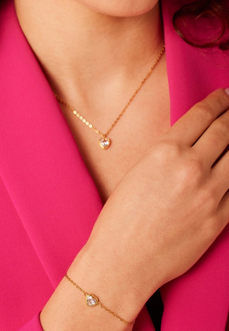 De gouden ketting en armband hebben hartvormige bedels met heldere stenen. Ze worden gedragen op een roze blouse. De ketting heeft een verfijnd ontwerp.