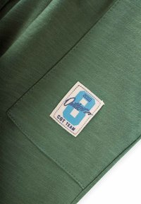 Grüner Stoff mit einer glatten Textur hat einen rechteckigen weißen Fleck. Der Fleck zeigt die blaue Zahl "6" und den Text "Outdoors COT TEAM."
