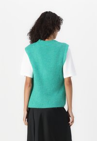 Knitted sleeveless top in teal, featuring a ribbed texture, round neckline, and relaxed fit, paired with a white short-sleeve shirt underneath.