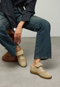 Beige suede sneakers with a foldable cuff, perforated design, and white laces. Paired with slightly flared denim jeans.