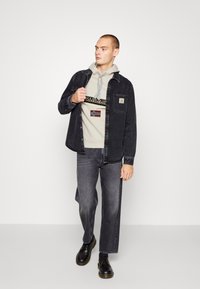 Camicia di jeans nera sopra un hoodie grigio chiaro con un logo, abbinata a jeans grigi e scarpe nere con lacci. Modello in un contesto neutro.