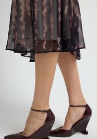 Tacones de cuña de charol burdeos con tira en el tobillo, que presentan una forma de cuña geométrica y un acabado brillante; combinados con un vestido fluido y estampado.