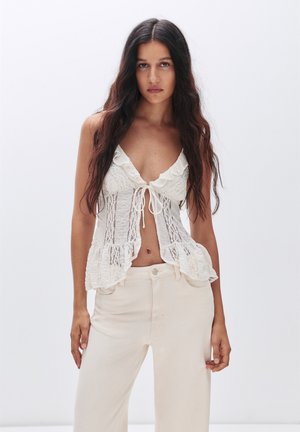 Hvid blondt cropped top med bølgede kanter, der har en knaplukning foran og gennemsigtige paneler. Sammen med lysebeige brettebukser.
