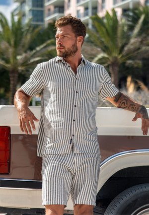 Mand med skæg og tatoveringer iført stribet kortærmet skjorte og matchende shorts læner sig op ad en hvid og brun lastbil udendørs.