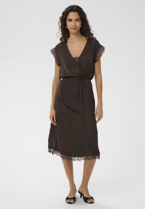 Žena s kučeravými vlasmi nosiaca tmavohnedé midi zavinovacie šaty s čipkovým lemom a čierne otvorené nízke podpätky stojaca na bielom pozadí.