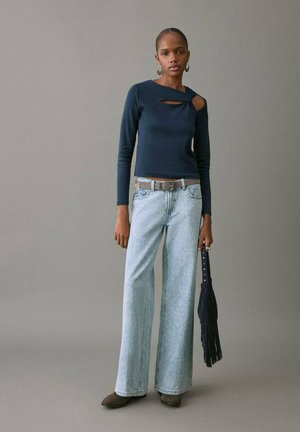 Top cu mâneci lungi, de culoare bleumarin, cu detaliu decupat, combinat cu blugi evazați deschiși la culoare. Fără buzunare, accesorizat cu o geantă bleumarin cu franjuri. Cizme maro de gleznă.