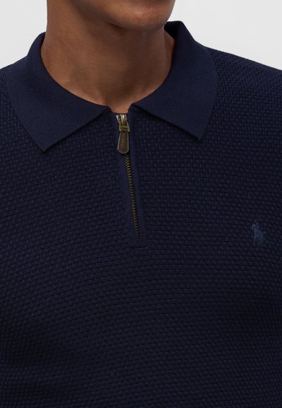 Homme portant un polo en maille texturée bleu marine avec une fermeture éclair dorée partiellement zippée et un col.