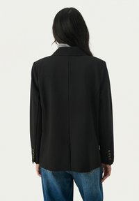 Čierny blazer so štruktúrovanou siluetou, s jedným zadným rozventilom a zlatými gombíkovými akcentmi na rukávoch. Tkanina vyzerá hladko s priliehavým strihom.