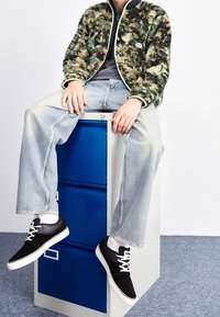 Flauschige Jacke mit grün-braunem Muster und weißen Besätzen, graues Shirt, hellblaue Jeans und schwarze Schuhe mit weißen Sohlen, sitzt auf einem blauen Aktenregal.