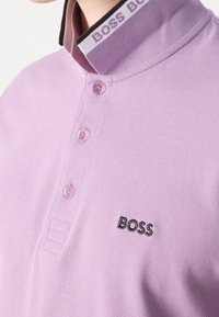 Polo lavande à trois boutons, logo noir "BOSS" sur la poitrine, col avec le texte "BOSS BOSS" en rayures noires et blanches.