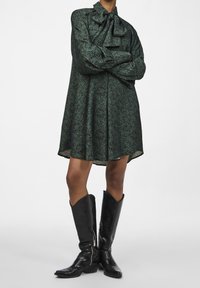 Green patterned dress with a bow neckline, long sleeves, and a relaxed fit, paired with knee-high black leather boots with zippers.